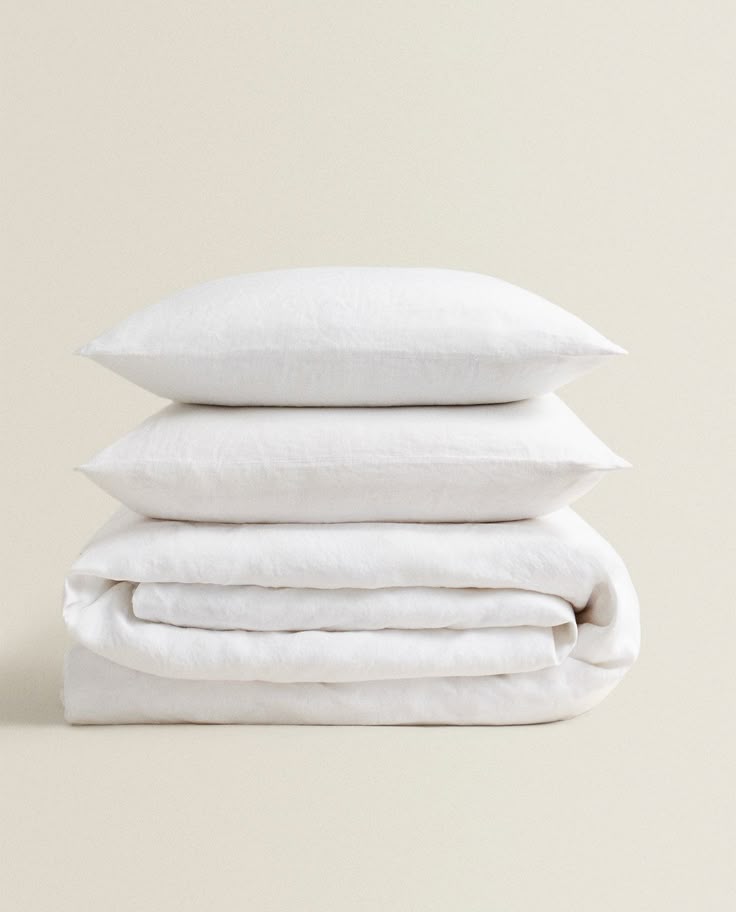 Two white pillows stacked on top of a folded white bedding set against a solid beige background.